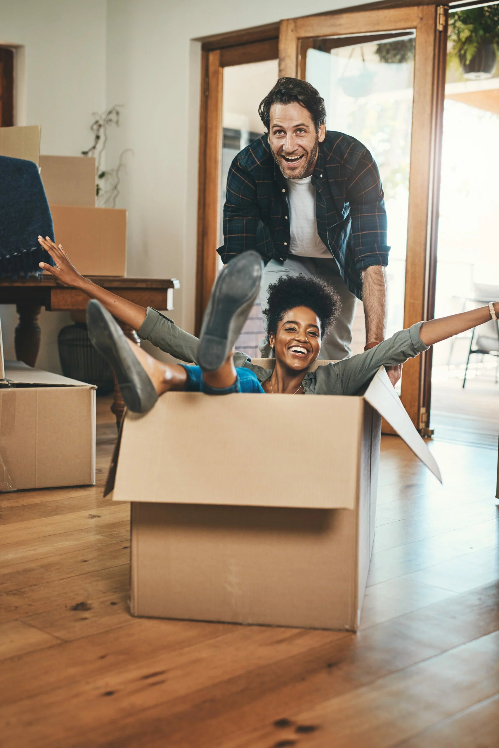 fun playful laughing homeowners playing box enjoying new home as real estate investors silly interracial couple happy man pushing woman living room purchased property scaled